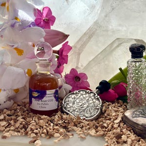 May include: A collection of vintage items, including a small glass bottle, a silver compact, and a perfume bottle, arranged with white and purple flowers. The scene is set on a bed of small pebbles.