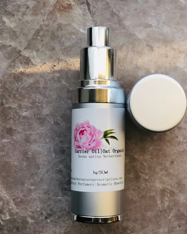 May include: A silver and white bottle of Carrier Oil Oat Organic with a pink peony illustration. The bottle is labeled with the text "Avena sativa Netherlands" and "1oz/30ml". The cap is off and placed to the side.