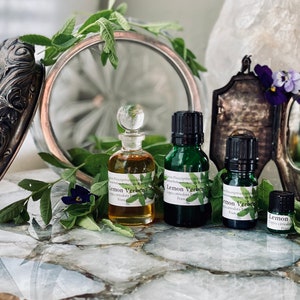 May include: An arrangement featuring antique silver-toned compacts and perfume bottles. The bottles, labeled "Lemon Verbena," are filled with amber and green liquids. Fresh green leaves and purple flowers are scattered around the bottles.
