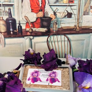 May include: A metal tin containing a light brown powder, surrounded by deep purple iris flowers. A label on the tin reads "Essential Oil", "Orris Root", and "Aged Iris x German". The background features a vintage illustration of a man in a pharmacy.