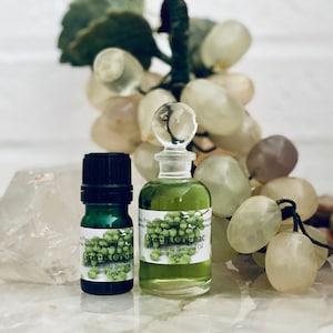 Könnte beinhalten: Zwei Flaschen Green Cognac ätherisches Öl, eine kleine und eine größere, mit Etiketten mit grünen Traubenillustrationen. Die Flaschen sind mit einer grünen Flüssigkeit gefüllt und vor dem Hintergrund einer dekorativen Traube angeordnet.