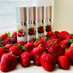 May include: Four silver-topped bottles of strawberry serum are surrounded by fresh, ripe strawberries. The bottles have labels with strawberry illustrations and text. The strawberries are a vibrant red with green leaves.