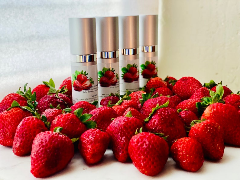 May include: Four silver-topped bottles of strawberry serum are surrounded by fresh, ripe strawberries. The bottles have labels with strawberry illustrations and text. The strawberries are a vibrant red with green leaves.