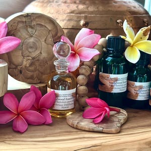 May include: An arrangement of essential oil bottles, a decorative glass bottle, wooden beads, and pink and yellow plumeria flowers. The bottles have black caps and labels. The background includes a copper pot and wooden accents.
