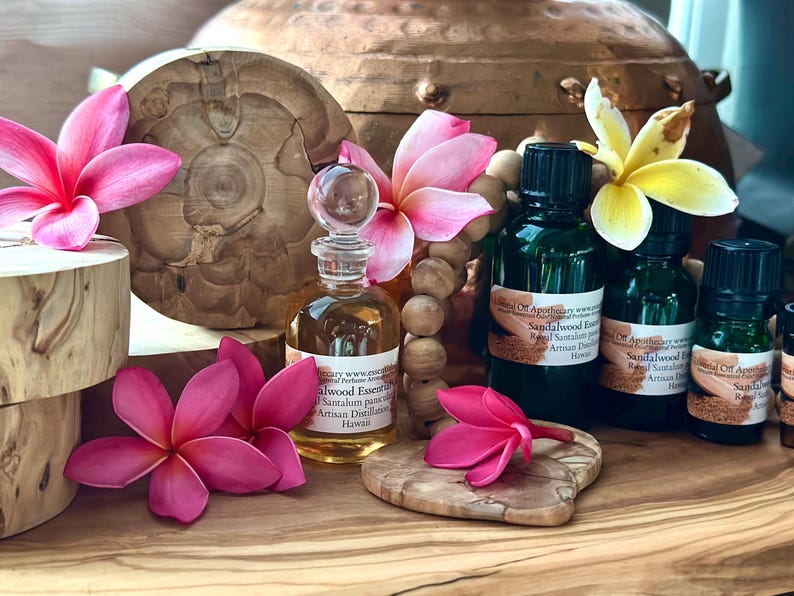May include: An arrangement of essential oil bottles, a decorative glass bottle, wooden beads, and pink and yellow plumeria flowers. The bottles have black caps and labels. The background includes a copper pot and wooden accents.