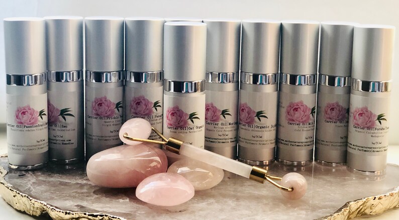May include: A collection of silver-topped bottles with labels featuring a pink peony design. A rose quartz face roller and several rose quartz stones are displayed in front of the bottles. The bottles are for carrier oils.