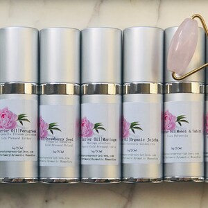 May include: An assortment of silver-topped bottles with labels featuring a pink peony design, alongside a rose quartz face roller with a gold handle. The bottles contain various carrier oils, each labeled with its botanical name.