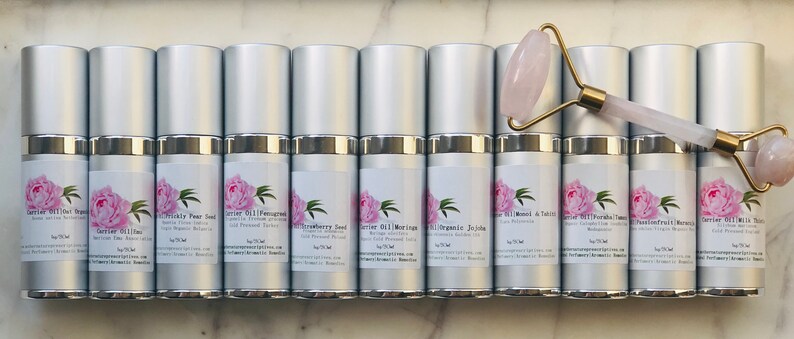 May include: An assortment of silver-topped bottles with labels featuring a pink peony design, alongside a rose quartz face roller with a gold handle. The bottles contain various carrier oils, each labeled with its botanical name.