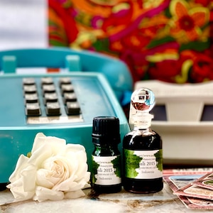 May include: Two small glass bottles of Patchouli essential oil with black caps and labels, beside a white rose and a vintage turquoise telephone. The background features a colourful floral pattern.