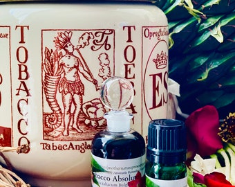 Può includere: Natura morta con un barattolo di tabacco vintage con coperchio dorato, una bottiglia di vetro e una piccola bottiglia verde. Il barattolo ha un'illustrazione marrone di un nativo americano che fuma la pipa. Le bottiglie contengono un liquido scuro e riportano la scritta "Tobacco Absolute".