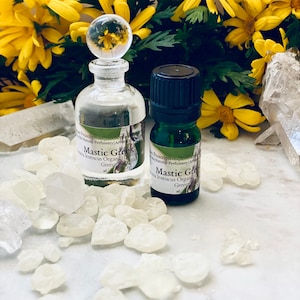 Könnte beinhalten: Zwei Glasflaschen mit ätherischem Öl, eine mit einem durchsichtigen Verschluss und die andere mit einer schwarzen Kappe, sind von weißen Kristallen und gelben Blumen umgeben. Die Flaschen sind mit "Mastic Greek" und "Mastic Gro" beschriftet.