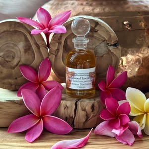 May include: A small glass bottle of sandalwood essential oil with a clear stopper, surrounded by pink and yellow plumeria flowers. The bottle sits on a wooden block, with a wooden background and a copper pot in the background.