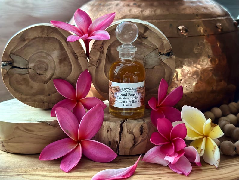 May include: A small glass bottle of sandalwood essential oil with a clear stopper, surrounded by pink and yellow plumeria flowers. The bottle sits on a wooden block, with a wooden background and a copper pot in the background.