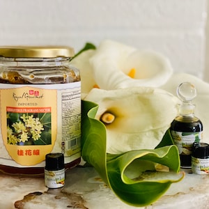 May include: A jar of Royal Gourmet Osmanthus Fragrans Nectar with a gold lid, alongside small bottles of Osmanthus perfume. White calla lilies and green leaves are in the background. The jar label includes Chinese characters and the words "Imported" and "Nectar".