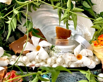 May include: A small glass jar of Jasmine Floral Wax with a silver lid, surrounded by white flowers, pearls, and brown wax pieces. The jar has a label with text. Green leaves and orange flowers frame the scene.