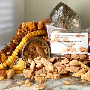 Puede incluir: Una variedad de productos aromáticos naturales, que incluyen joyas de ámbar, un frasco de vidrio lleno de trozos de resina, una botella de líquido oscuro y un paquete etiquetado como "Premium Styrax Benzoin 'Almonds' Resin Thailand."