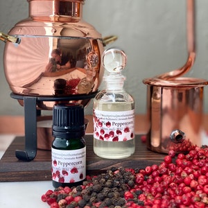 May include: A copper still with a small glass bottle labeled "Pink Peppercorn" and a dark green bottle labeled "Peppercorn." Red and black peppercorns are scattered on a wooden surface. The still is on a black metal stand.