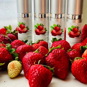 May include: Four silver-topped bottles of strawberry seed oil are displayed with a backdrop of fresh, ripe strawberries. The bottles have labels with strawberry illustrations and text. The strawberries are a vibrant red.