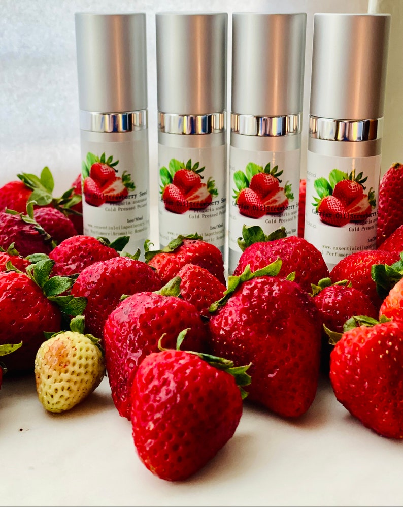 May include: Four silver-topped bottles of strawberry seed oil are displayed with a backdrop of fresh, ripe strawberries. The bottles have labels with strawberry illustrations and text. The strawberries are a vibrant red.