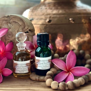 May include: Two small glass bottles of sandalwood essential oil with text, surrounded by pink flowers and wooden beads. One bottle is clear with a glass stopper, the other is dark green. The text reads "Sandalwood Essential Oil, Artisan Distillation, Hawaii."