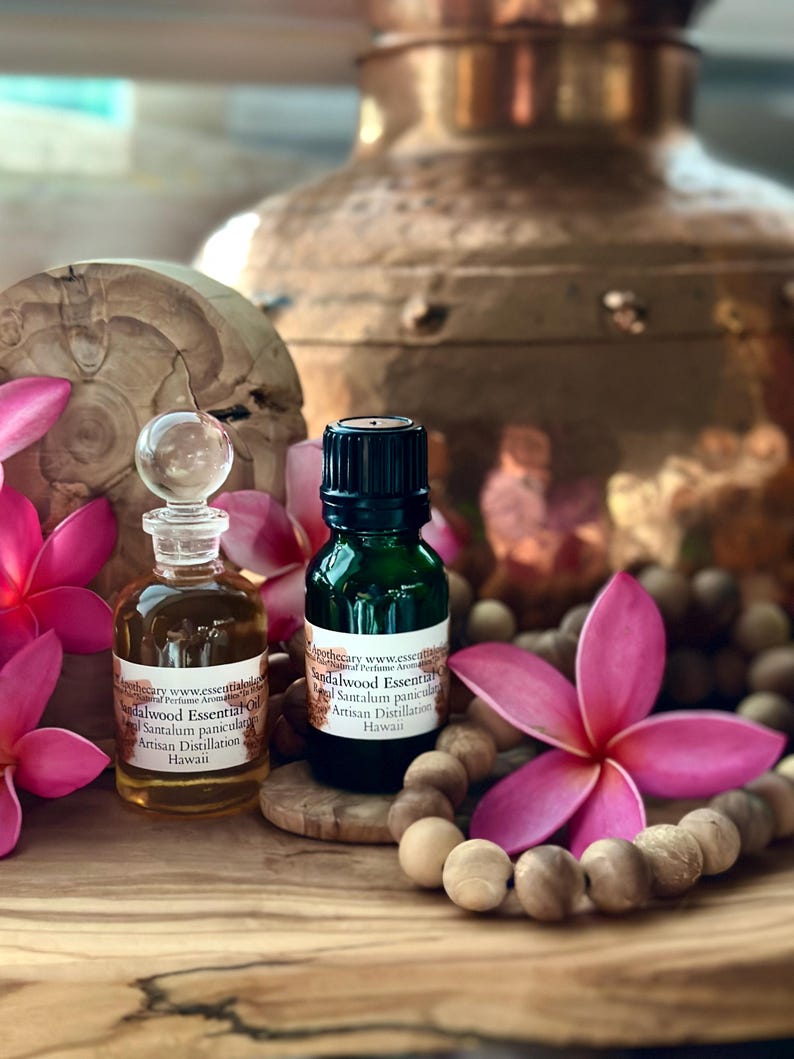 May include: Two small glass bottles of sandalwood essential oil with text, surrounded by pink flowers and wooden beads. One bottle is clear with a glass stopper, the other is dark green. The text reads "Sandalwood Essential Oil, Artisan Distillation, Hawaii."