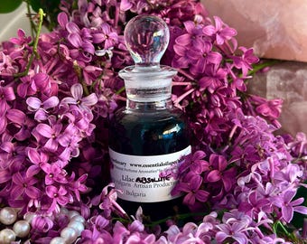 May include: A small, dark glass bottle with a clear glass stopper, surrounded by vibrant purple lilac blossoms. The bottle contains a dark liquid and has a label that reads "Lilac Absolute". A string of pearls and a pink crystal are also present.