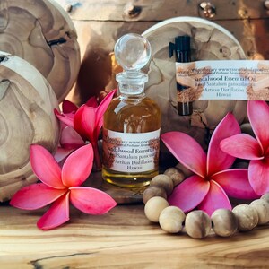May include: A clear glass bottle of sandalwood essential oil with a glass stopper, surrounded by pink flowers and a wooden bead necklace. The label reads "Sandalwood Essential Oil".