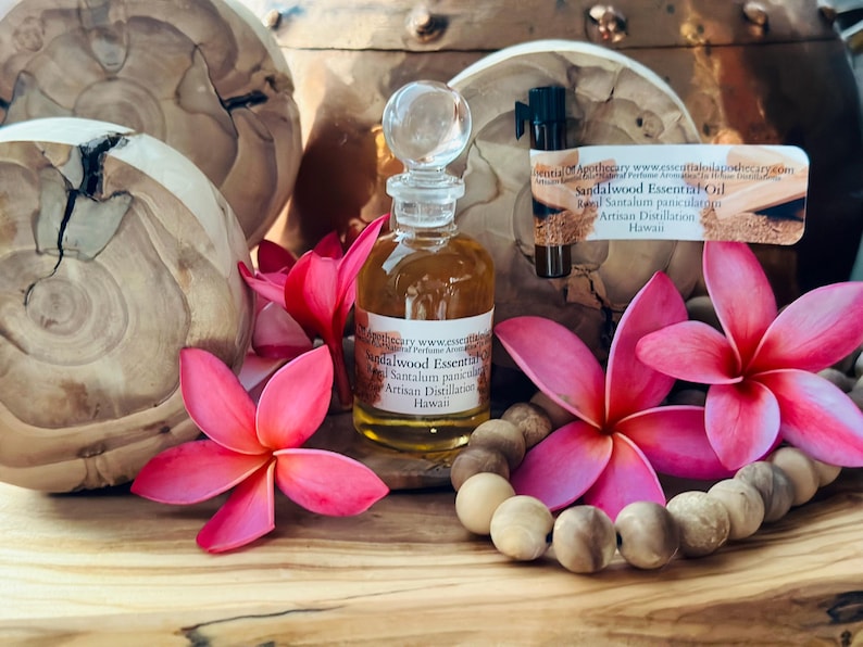 May include: A clear glass bottle of sandalwood essential oil with a glass stopper, surrounded by pink flowers and a wooden bead necklace. The label reads "Sandalwood Essential Oil".
