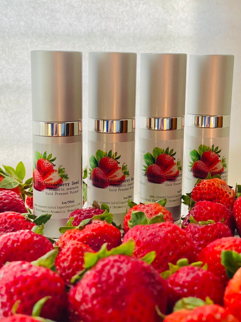 May include: Four silver and white essential oil bottles with labels featuring strawberries and text. The bottles are surrounded by fresh, ripe strawberries. The labels read "Strawberry Seed Fragaria ananassa Cold Pressed Poland 1oz/30ml".