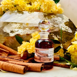 May include: A small, brown glass bottle with a clear glass stopper, labeled "Massoia Bark Essence." The bottle is surrounded by cinnamon sticks, yellow flowers, and a large, clear crystal. The label also includes the words "Essential Oils" and "Natural Perfumery."