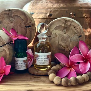 May include: A still life featuring essential oil bottles, wooden accents, and vibrant pink plumeria flowers. The scene includes a small green bottle and a clear glass bottle with a decorative stopper. A wooden bead garland and a copper vessel complete the composition.