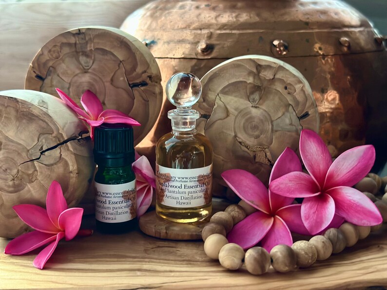 May include: A still life featuring essential oil bottles, wooden accents, and vibrant pink plumeria flowers. The scene includes a small green bottle and a clear glass bottle with a decorative stopper. A wooden bead garland and a copper vessel complete the composition.