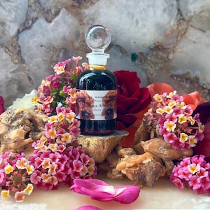 May include: A dark glass bottle with a clear stopper, surrounded by pink, yellow, and red flowers. The bottle sits on a gold-colored base, with a label visible. The background is a light-colored stone.