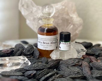 Può includere: Due piccole bottiglie di vetro di profumo Tonka Bean Absolut, una con tappo trasparente e l'altra con tappo nero, sono poste tra fagioli tonka neri e cristalli di quarzo trasparente. Le bottiglie hanno etichette con testo.