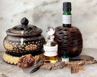 Può includere: Un assortimento di prodotti Attar of Oud, tra cui piccoli flaconi di vetro, un bruciatore di incenso nero e oro e pezzi di legno di oud. I flaconi hanno etichette trasparenti. Il bruciatore di incenso è nero con motivi floreali dorati.