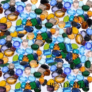 May include: Assortment of faceted gemstone beads in various shapes and colors, including blue, green, brown, and clear. The beads are scattered on a white background.