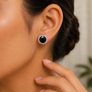 May include: A round stud earring featuring a faceted black stone set in a silver-toned metal frame with a twisted rope-style border. The earring is secured to the earlobe with a post back, showcasing a circular design with a polished finish.