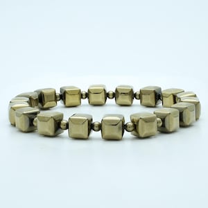 Puede incluir: Un brazalete dorado hecho de cuentas cuadradas. El brazalete está hecho de una serie de cuentas cuadradas conectadas.