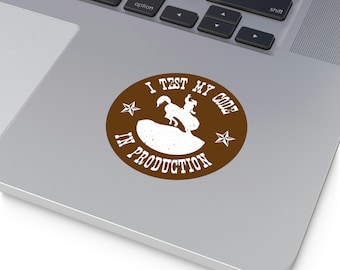 May include: A brown circle sticker with a white cowboy riding a bucking bronco. The sticker says "I test my code in production" with two white stars on either side of the text.