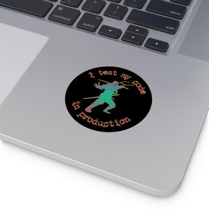 May include: A black and white sticker with a silhouette of a ninja holding a sword. The text "I test my code in production" is written in a rainbow gradient.