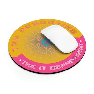 May include: A round pink and yellow mouse pad with the text "The IT Department is Booting Up" in a pixelated font. A white computer mouse is on top of the mouse pad.