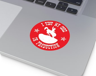 May include: Red circle sticker with white text that reads "I TEST MY CODE IN PRODUCTION". A white silhouette of a cowboy riding a bucking bronco is inside the circle. Two white stars are on either side of the cowboy.