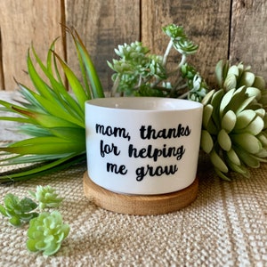 May include: White ceramic planter with a wooden base. The planter has black text that reads "mom, thanks for helping me grow".