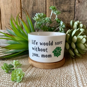 May include: White ceramic planter with a wooden base. The planter has the text "life would succ without you, mom" printed on it. There is a green succulent graphic on the right side of the planter.