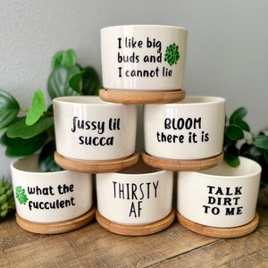 Puede incluir: Conjunto de seis macetas de suculentas de cerámica blanca con frases divertidas. Las macetas están apiladas en forma de pirámide y tienen bases de madera. Las frases en las macetas son: "I like big buds and I cannot lie", "what the fucculent", "fussy lil succa", "THIRSTY AF", "BLOOM there it is", y "TALK DIRT TO ME".