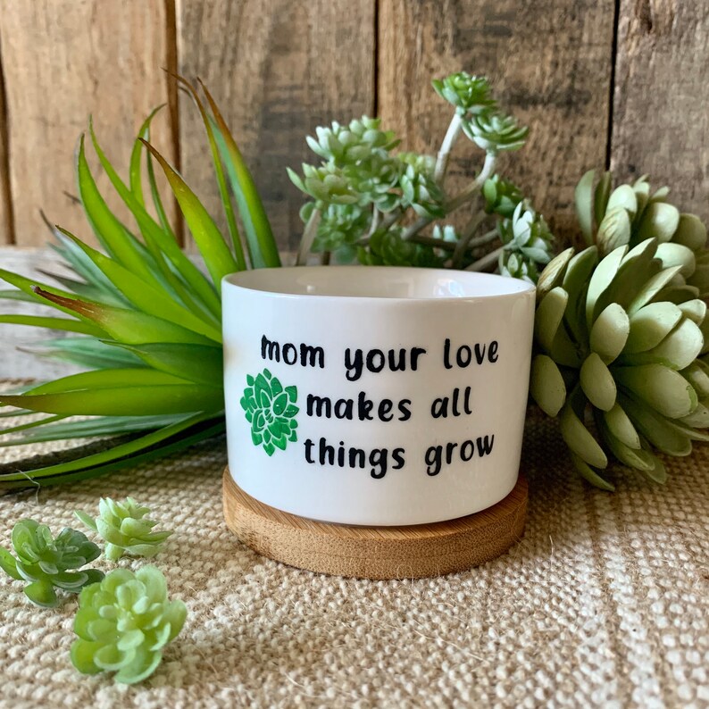 May include: White ceramic planter with a green succulent graphic and the text "Mom your love makes all things grow". The planter is sitting on a wooden coaster.