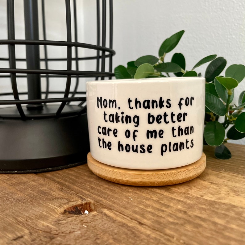 May include: White ceramic planter with a bamboo base. The planter has black text that reads "Mom, thanks for taking better care of me than the house plants."