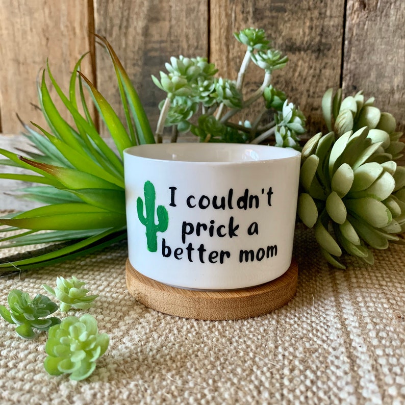 May include: White ceramic planter with a green cactus graphic and the text "I couldn't prick a better mom". The planter is sitting on a wooden coaster.