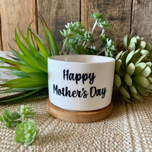 May include: White ceramic planter with the text "Happy Mother's Day" in black lettering. The planter is sitting on a wooden coaster.