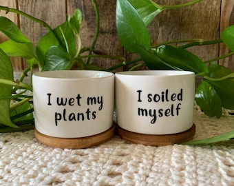 May include: Two white ceramic plant pots with bamboo bases. The pot on the left says "I wet my plants" and the pot on the right says "I soiled myself".
